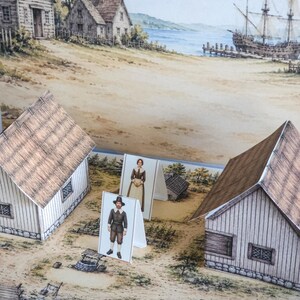 May include: A paper craft set featuring two three-dimensional houses and two standing figures of people in historical attire. The scene is set on a printed base depicting a coastal village with a well, wooden structures, and a large sailing ship.