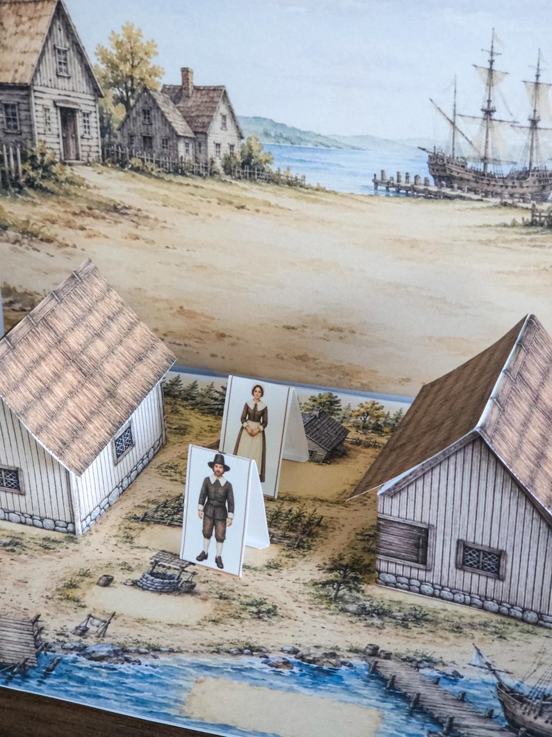 May include: A paper craft set featuring two three-dimensional houses and two standing figures of people in historical attire. The scene is set on a printed base depicting a coastal village with a well, wooden structures, and a large sailing ship.