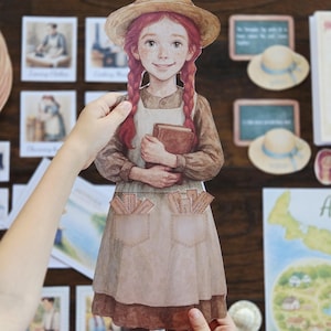 以下が含まれることがあります： 赤い三つ編みの女の子の紙の切り抜き。麦わら帽子とエプロンを着て、本を持っています。切り抜きは手で持たれており、背景には帽子や地図など、他の紙の切り抜きやイラストがあります。
