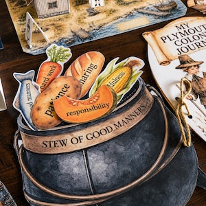 May include: A paper cutout of a dark gray cauldron labeled STEW OF GOOD MANNERS, filled with illustrated food items including a fish, carrot, potato, bread, corn, and pumpkin slice, each printed with words like patience and responsibility.