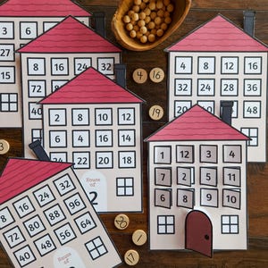 May include: A set of paper house-shaped educational cards with red roofs, each featuring a grid of numbered windows and a door. Some houses are labeled House of 2 or House of 8. Small wooden tokens with numbers are scattered near a wooden bowl of beads.