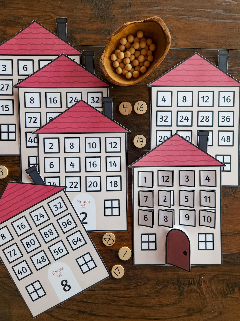 May include: A set of paper house-shaped educational cards with red roofs, each featuring a grid of numbered windows and a door. Some houses are labeled House of 2 or House of 8. Small wooden tokens with numbers are scattered near a wooden bowl of beads.