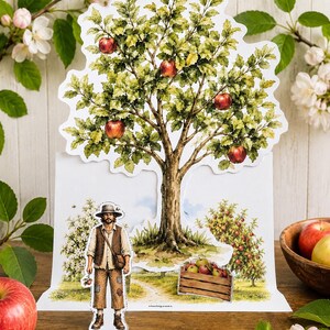 May include: A die-cut paper display featuring a large apple tree with green leaves and red apples. A separate paper cutout of a person wearing a hat, vest, and pants stands in front of a scene with a wooden crate filled with apples and a small orchard.