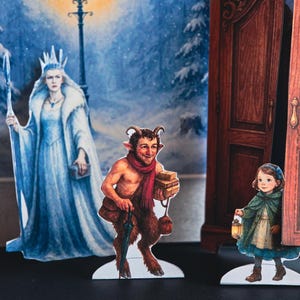 May include: A pop-up book scene featuring illustrated characters in a winter setting. The characters include a queen in a white gown, a faun with a red scarf, and a girl holding a lantern. A wardrobe and a lamppost are also visible.