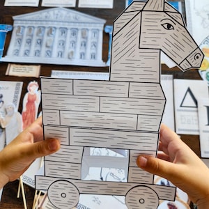 May include: A papercraft Trojan horse, held by hands, with a detailed design of wooden planks and wheels. The horse is gray and white, with a detailed head and eye. The background includes other educational materials.