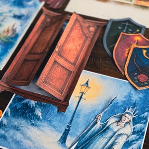 May include: A collection of fantasy-themed paper cutouts, including a wardrobe, shields, and illustrations of a winter scene with a queen and a lamppost. The color palette includes shades of brown, blue, and white.