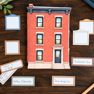 May include: A die-cut paper building shaped like a three-storey red brick townhouse with black window frames and a front door. Several rectangular paper labels with handwritten text including Wilbur is danger, She writes her web, Wilbur & Charlotte, Zuckerman's farm, and Spring through Fall are arranged around the building.