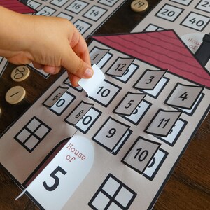 May include: An educational math game featuring paper houses with red roofs and windows that open to reveal numbers. Each house is labeled House of 5, House of 10, or similar. Small wooden circular tokens with printed numbers are scattered around the game boards.