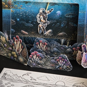 May include: A multi-layered paper art piece depicting an underwater scene with an astronaut. The scene includes coral reefs, marine life, and an astronaut figure. The artwork has a detailed, colorful design with a pop-up effect, creating a 3D appearance.