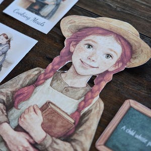 以下が含まれることがあります： 赤い三つ編みと麦わら帽子をかぶった女の子の切り絵。本を手に持っています。彼女の周りには、「Cooking Meals」やテキストが書かれた小さな黒板など、イラストとテキストが描かれた他の切り絵があります。
