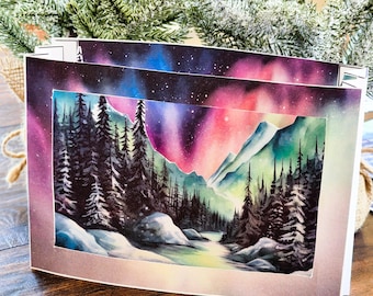 May include: A set of two greeting cards featuring a watercolor painting of a snowy forest scene with a river flowing through it. The sky is a vibrant pink, purple, and blue with stars. The cards are perfect for sending a wintery greeting.