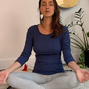 May include: A woman sits in a meditative pose on a black and white patterned yoga mat with her eyes closed. She is wearing a blue long-sleeved shirt and grey leggings. The mat is on a white rug.