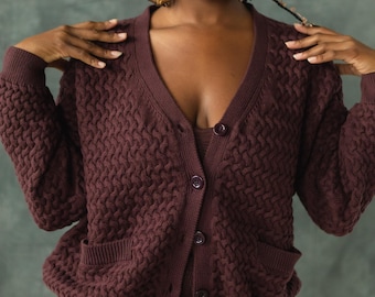 May include: A woman wearing a brown knitted cardigan with a cable knit pattern and a v-neck. The cardigan has two pockets and is buttoned up the front.