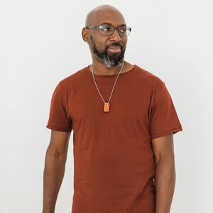 Pu&ograve; includere: Un uomo che indossa una maglietta marrone con una collana a catena d'argento e un piccolo ciondolo rettangolare. Indossa pantaloni arancioni e ha la testa rasata e gli occhiali.