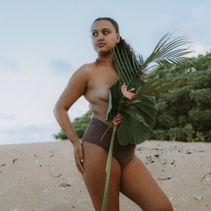 May include: A person standing on a sandy beach wearing brown high-waisted underwear and holding a large green leaf with a pink flower.