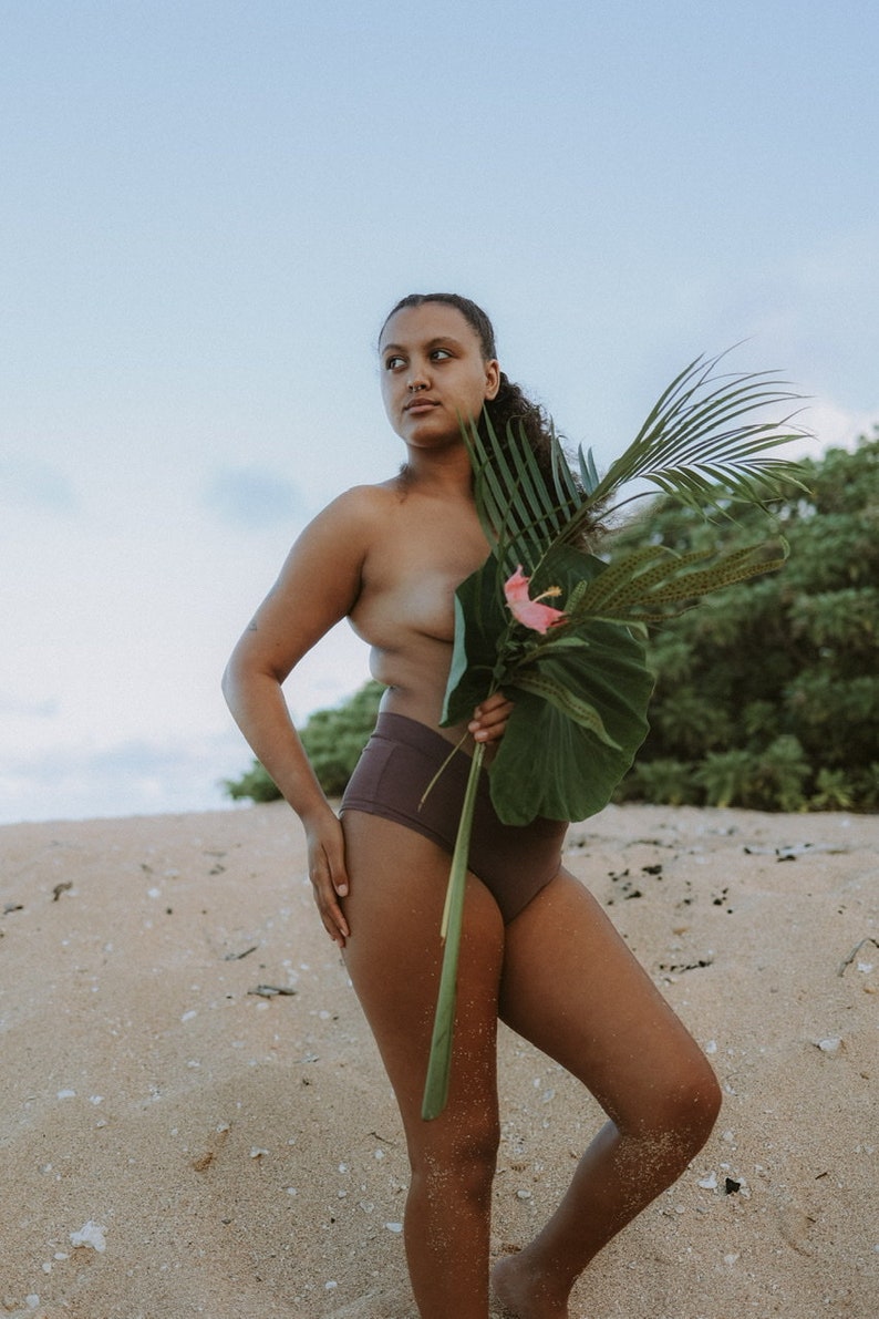 May include: A person standing on a sandy beach wearing brown high-waisted underwear and holding a large green leaf with a pink flower.