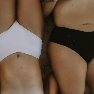 May include: Two people are lying down on a sandy surface, wearing swimwear. One is wearing a white bikini bottom, and the other is wearing a black bikini bottom. The image is taken from a high angle.