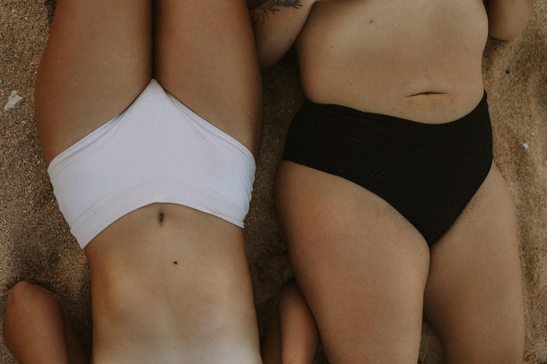 May include: Two people are lying down on a sandy surface, wearing swimwear. One is wearing a white bikini bottom, and the other is wearing a black bikini bottom. The image is taken from a high angle.
