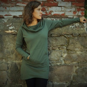 Könnte beinhalten: Eine Frau trägt ein dunkelgrünes Kapuzenpulloverkleid mit einer großen Tasche auf der Vorderseite. Sie steht vor einer Steinmauer.