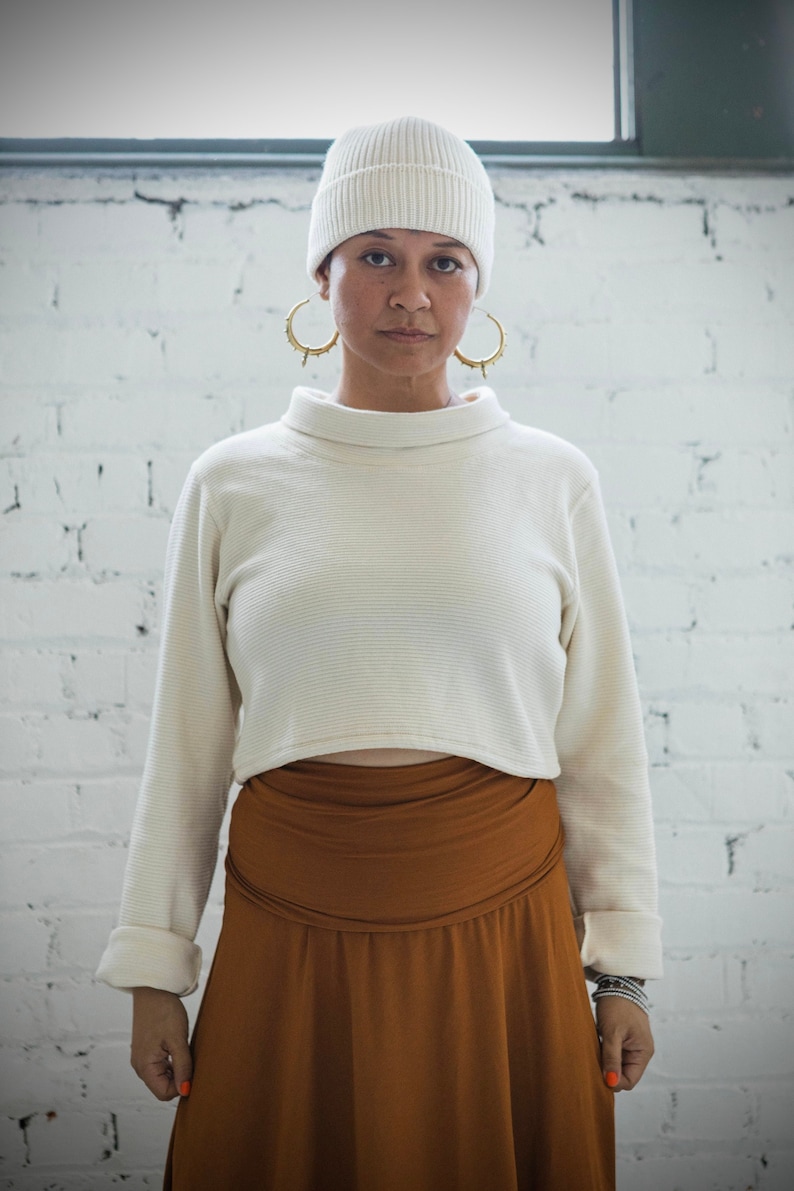 May include: A woman wearing a white knit beanie, a white long-sleeved sweater, and a brown skirt. She is standing in front of a white brick wall.