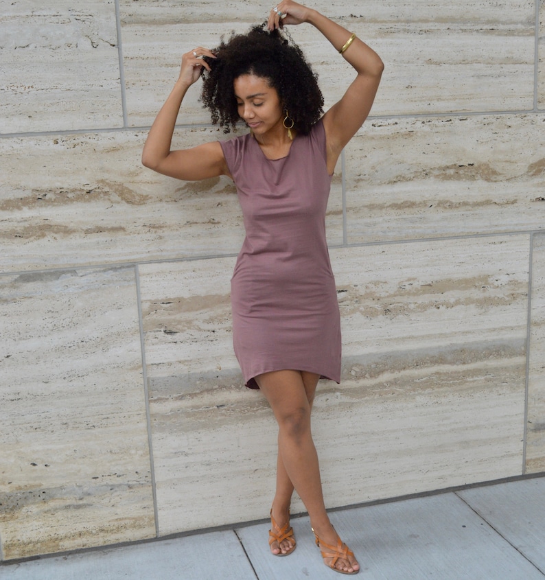 May include: A mauve-colored sleeveless dress featuring a round neckline and a hemline that is slightly longer in the back than in the front. The garment is constructed from a solid, smooth fabric and falls to a length above the knees.