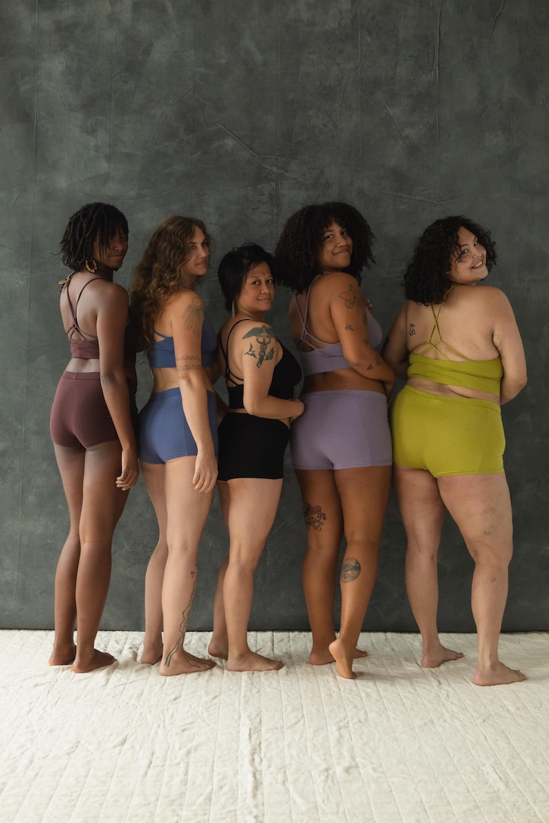 May include: Five people stand in a row, facing away from the camera, wearing different colored underwear. The underwear is all the same style, with a high waist and a wide leg.