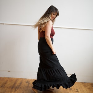 May include: A woman wearing a maroon tank top and a black maxi skirt with ruffles. She is standing on a wooden floor and is twirling the skirt.