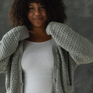 May include: A woman wearing a gray knitted cardigan with a cable knit pattern and a white tank top. The cardigan has a button closure and pockets.
