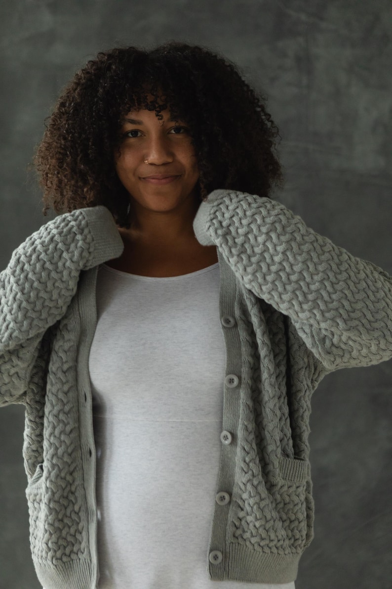 May include: A woman wearing a gray knitted cardigan with a cable knit pattern and a white tank top. The cardigan has a button closure and pockets.