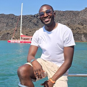 Pu&ograve; includere: Un uomo con occhiali da sole e una maglietta bianca &egrave; seduto sul corrimano di una barca. Indossa pantaloncini beige e un orologio. La barca &egrave; in acqua con una barca rossa sullo sfondo. Il testo "sunset-oia.com" &egrave; visibile sulla barca rossa.