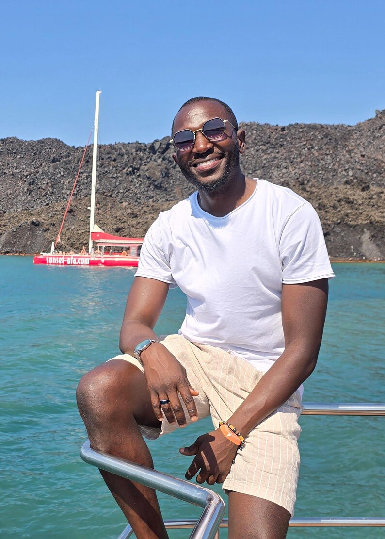 Pu&ograve; includere: Un uomo con occhiali da sole e una maglietta bianca &egrave; seduto sul corrimano di una barca. Indossa pantaloncini beige e un orologio. La barca &egrave; in acqua con una barca rossa sullo sfondo. Il testo "sunset-oia.com" &egrave; visibile sulla barca rossa.