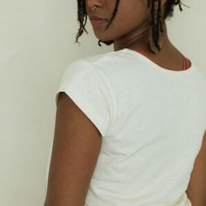 May include: Cream-colored short-sleeved t-shirt. The shirt has a rounded neckline and rolled-up sleeves. The model is wearing a rust-colored top underneath and mustard-colored pants. The model has dark hair and is looking over their shoulder.