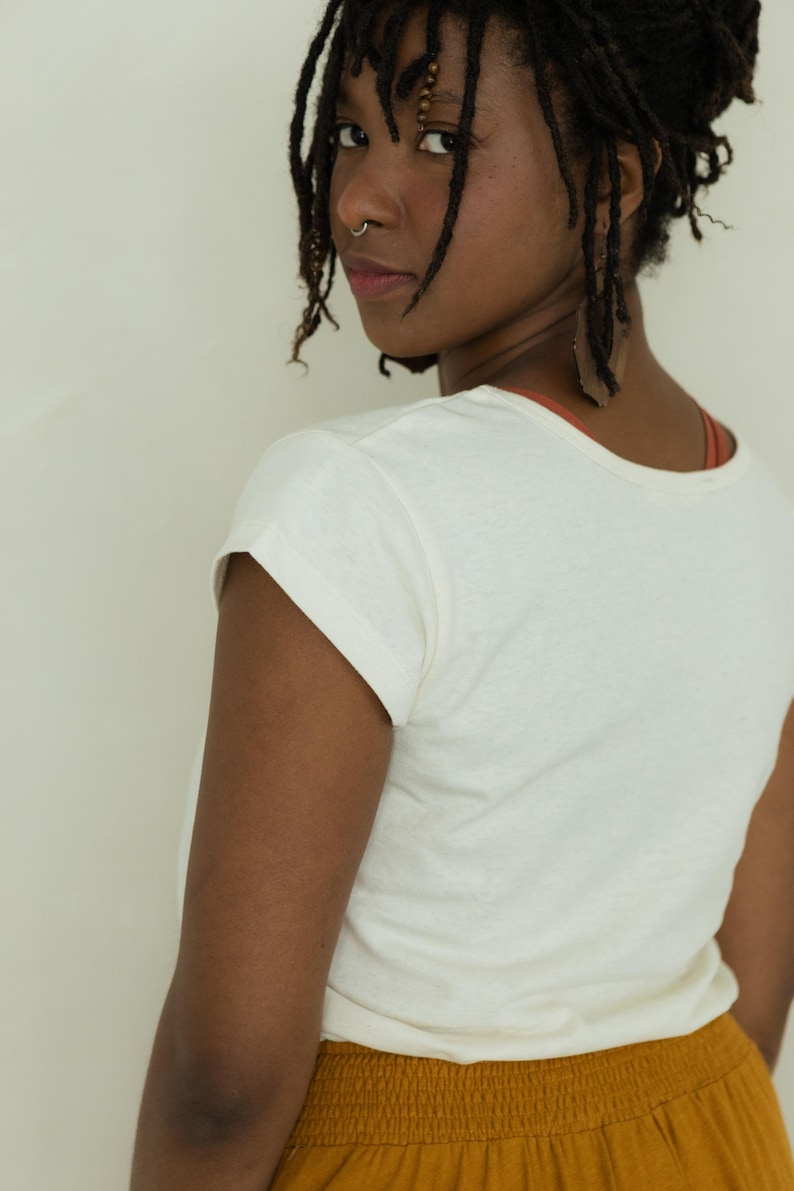 May include: Cream-colored short-sleeved t-shirt. The shirt has a rounded neckline and rolled-up sleeves. The model is wearing a rust-colored top underneath and mustard-colored pants. The model has dark hair and is looking over their shoulder.