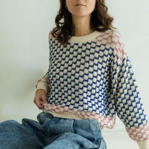 May include: A cream-colored sweater with a blue and white checkered pattern. The sweater has pink accents on the neckline and cuffs. The model is wearing acid wash blue jeans.