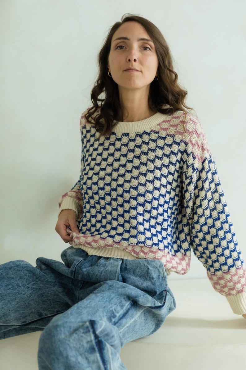 May include: A cream-colored sweater with a blue and white checkered pattern. The sweater has pink accents on the neckline and cuffs. The model is wearing acid wash blue jeans.