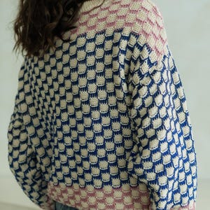 May include: A knitted sweater with a unique pattern. The sweater features a repeating geometric design in navy blue, cream, and pink. The sleeves and neckline are finished with a ribbed trim. The overall style is casual and comfortable.