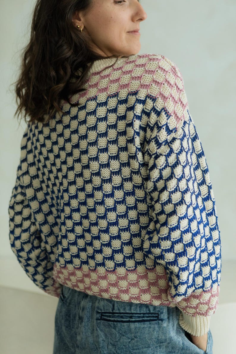 May include: A knitted sweater with a unique pattern. The sweater features a repeating geometric design in navy blue, cream, and pink. The sleeves and neckline are finished with a ribbed trim. The overall style is casual and comfortable.