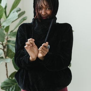 May include: A black hooded sweatshirt with a high collar and drawstring. The sweatshirt is made of a soft, velvety material. The person is wearing burgundy leggings. A large green plant is in the background.