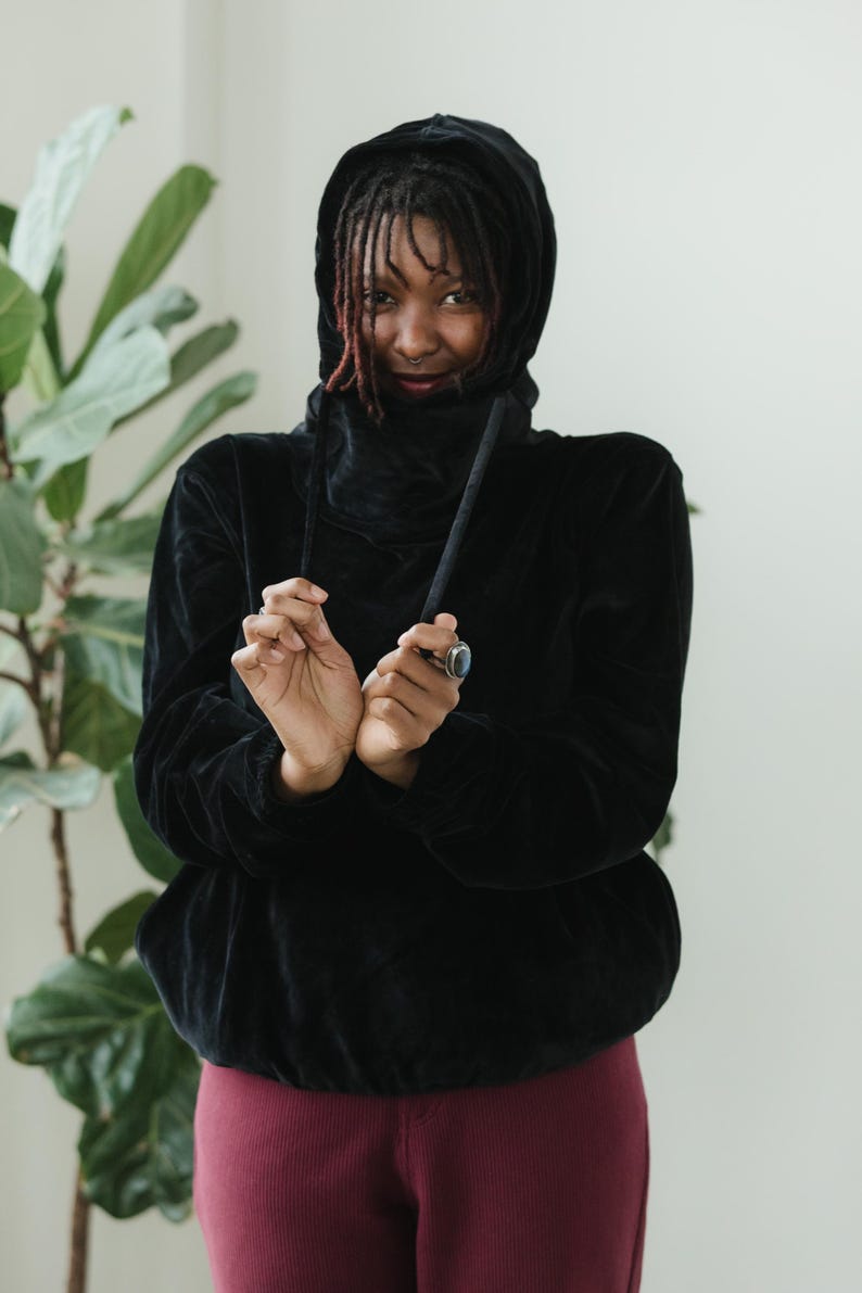 May include: A black hooded sweatshirt with a high collar and drawstring. The sweatshirt is made of a soft, velvety material. The person is wearing burgundy leggings. A large green plant is in the background.