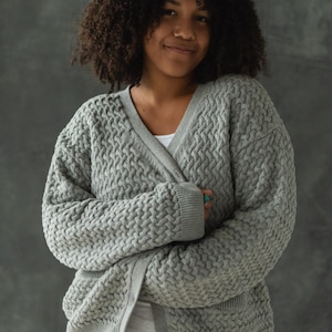 May include: A woman wearing a gray cable knit cardigan with a v-neck and button closure. The cardigan has a textured pattern and is worn over a white shirt.