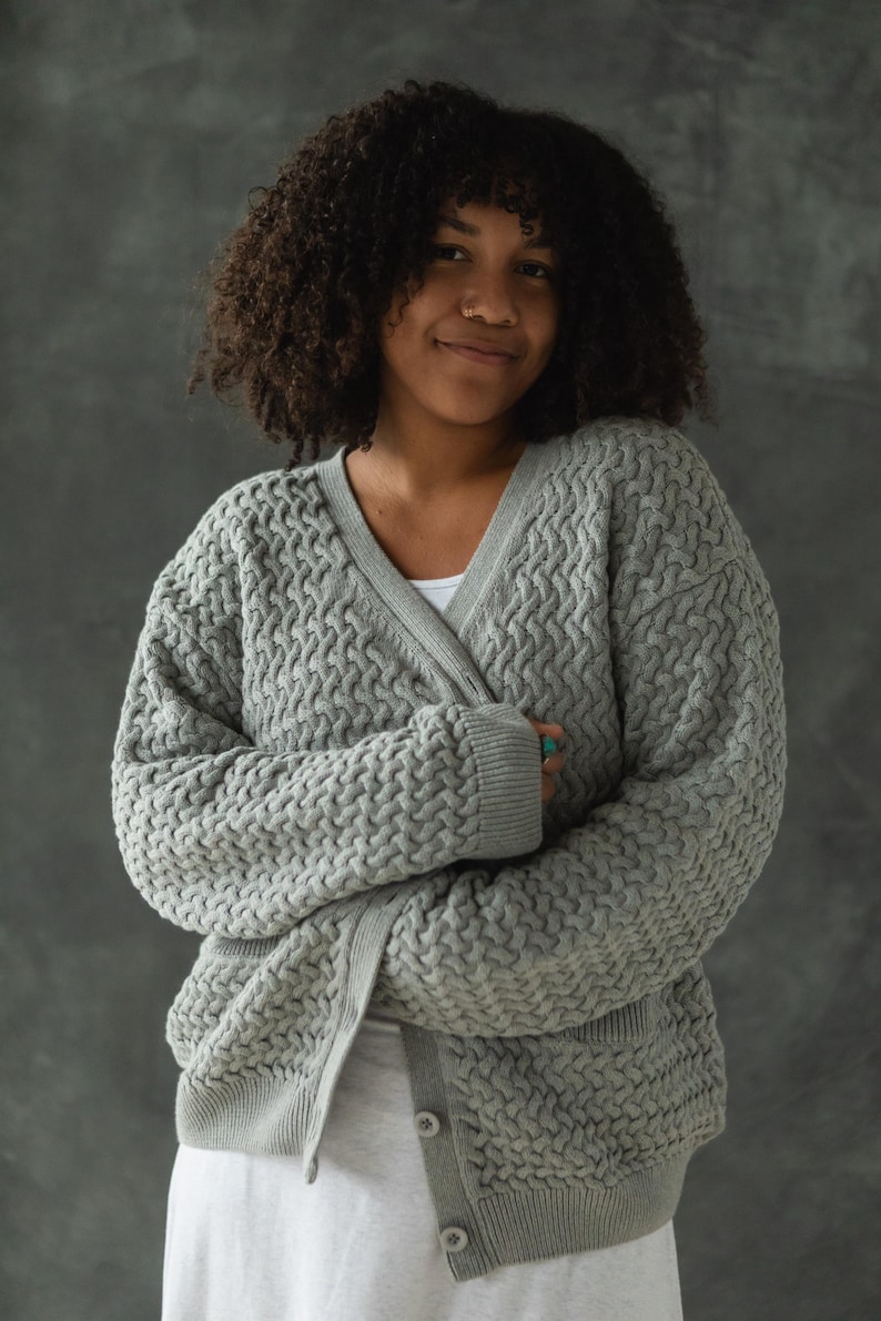 May include: A woman wearing a gray cable knit cardigan with a v-neck and button closure. The cardigan has a textured pattern and is worn over a white shirt.