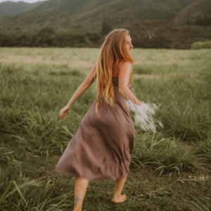 May include: A solid mauve-colored midi dress with a straight neckline and thin straps. The garment features a simple, fluid silhouette that falls below the knees, crafted from a smooth, lightweight fabric that allows for movement while walking.