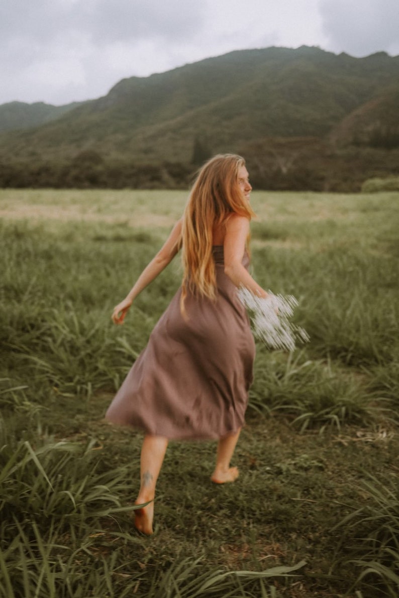May include: A solid mauve-colored midi dress with a straight neckline and thin straps. The garment features a simple, fluid silhouette that falls below the knees, crafted from a smooth, lightweight fabric that allows for movement while walking.