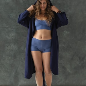 May include: A woman wearing a dark blue robe with a hood, a light blue bra, and light blue shorts. She is standing in front of a gray textured background.