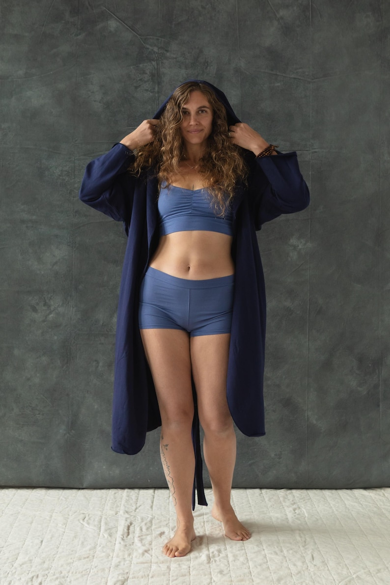 May include: A woman wearing a dark blue robe with a hood, a light blue bra, and light blue shorts. She is standing in front of a gray textured background.