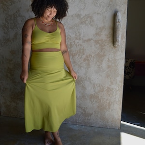 May include: A two-piece set consisting of a chartreuse crop top with thin spaghetti straps and a ruched center detail, paired with a matching high-waisted, floor-length skirt. The fabric is a solid, smooth material in a vibrant yellow-green hue.