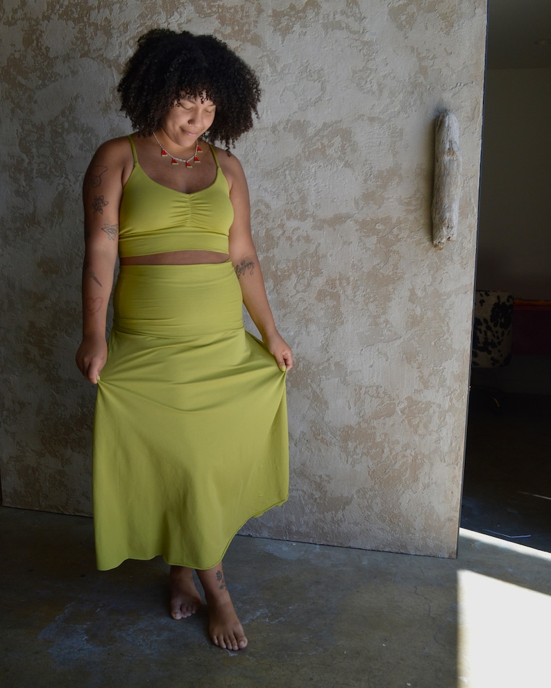 May include: A two-piece set consisting of a chartreuse crop top with thin spaghetti straps and a ruched center detail, paired with a matching high-waisted, floor-length skirt. The fabric is a solid, smooth material in a vibrant yellow-green hue.