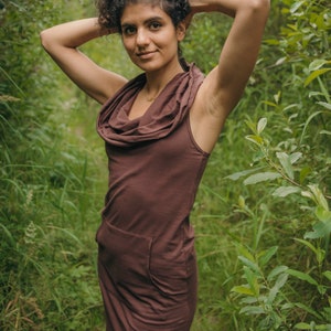 May include: A woman wearing a brown cowl neck dress with a single pocket on the left side. She is standing in a forest setting with green foliage.
