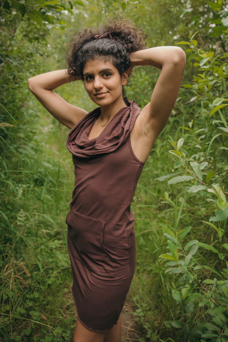 May include: A woman wearing a brown cowl neck dress with a single pocket on the left side. She is standing in a forest setting with green foliage.