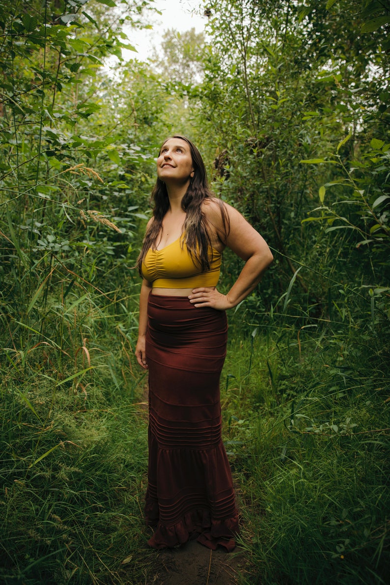 May include: A woman wearing a yellow crop top and a long, burgundy skirt stands in a lush green forest. She is looking up at the sky with a thoughtful expression.